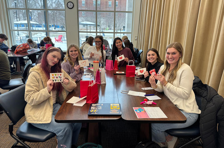 A group of six people sit around a rectangular table in a bright room with large windows. They are smiling and holding up small craft items or cards. Red gift bags, markers, and papers are spread across the table. Snow can be seen outside the windows.