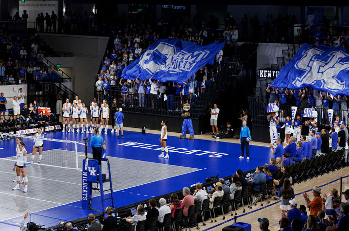  Fans hold large blue Kentucky Wildcats flags in the stands as UK volleyball players stand on the court.