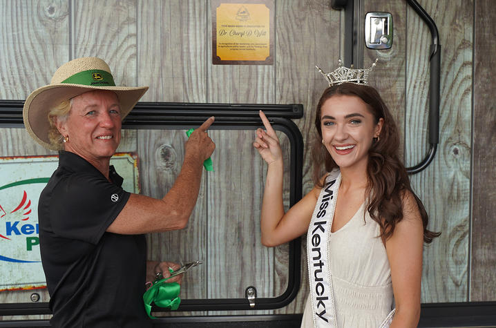 Two people stand in front of a wooden wall and point upward toward a mounted plaque. One wears a wide-brimmed hat and gloves, while the other wears a sash and a decorative headpiece.
