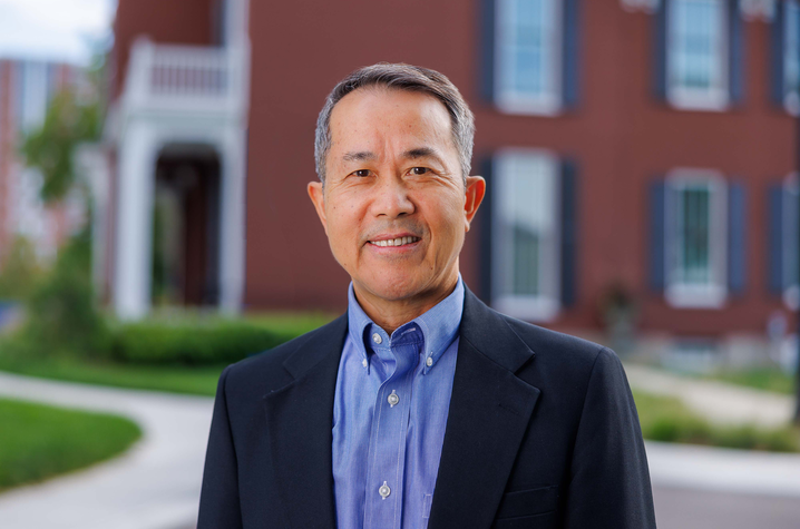 Man standing in front of a red brick building, smiling for a portrait, dressed in a dark blazer and blue shirt