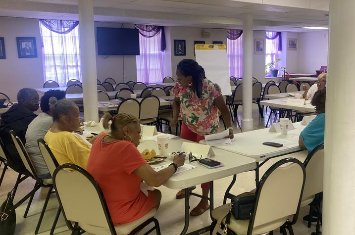 A focus group meets to discuss health information handouts. Eleven Black adults sit at long folding tables arranged in a U-shape. In the center, a Black woman leading the group leans over one of the tables to talk to a participant. 