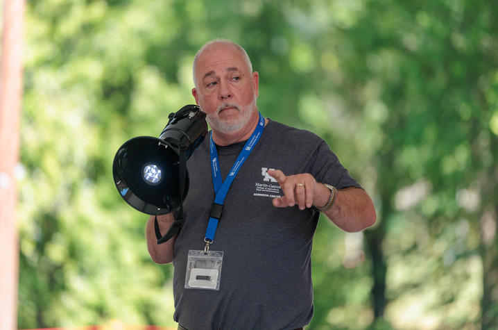 A presenter wearing a lanyard and name badge speaks to an audience while holding a handheld megaphone under an outdoor pavilion. The presenter gestures with one hand, with trees and greenery blurred in the background.