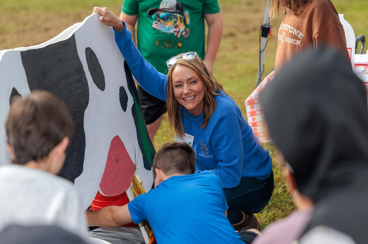 An educator kneels beside a large cow display while leading a dairy science activity for a group of children outdoors. Several children gather around the demonstration, with one child interacting with the display as the instructor explains the activity.