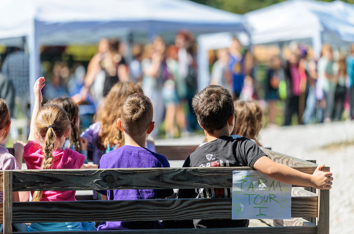 Several children sit on a wooden wagon during a farm tour, facing a crowd gathered near white canopy tents in the background. A handwritten sign on the wagon reads “Farm Tour 1,” and one child raises a hand while the group waits in an outdoor event area.