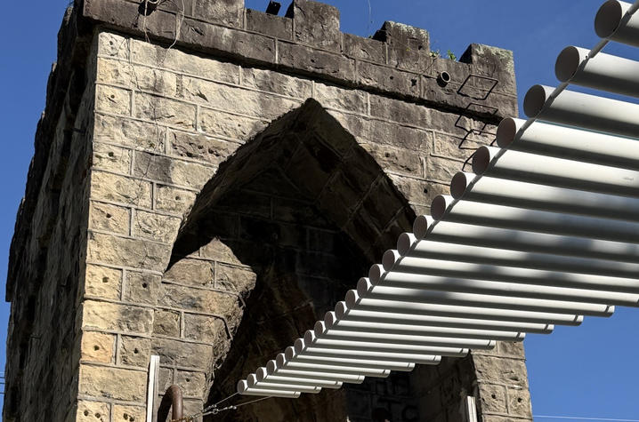 A historic stone bridge tower stands beneath a clear blue sky, with a modern white overhead canopy extending from the structure. Sunlight casts striped shadows across the stone façade, highlighting the contrast between the old masonry and new design.