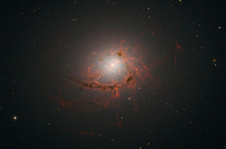 Image of a glowing elliptical galaxy surrounded by reddish filaments of gas, with a bright central core against a dark, star-filled background.