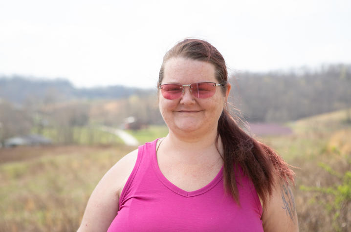 a headshot of manning standing with a beautiful eastern kentucky landscape in the background. She's wearing a pink sleeveless top and has matching pink sunglasses on. she's smiling.