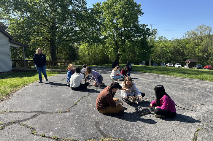 People sit and kneel on a paved outdoor area, working together on an activity with papers and materials spread on the ground near a building and trees.