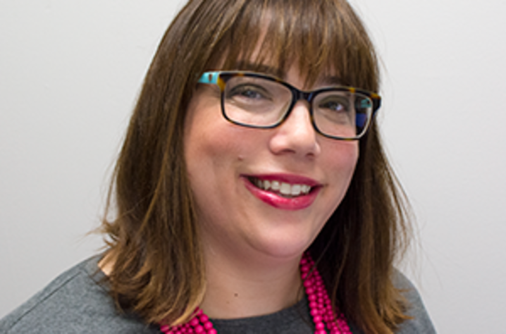 Headshot of Heather M. Bush, Ph.D.. She's in front of a white wall and wears a gray shirt, a multi-stranded necklace of pink peral-shaped beads, black glasses, and red lipstick. She medium-brown hair down to her should and hangs. She's smiling at the came