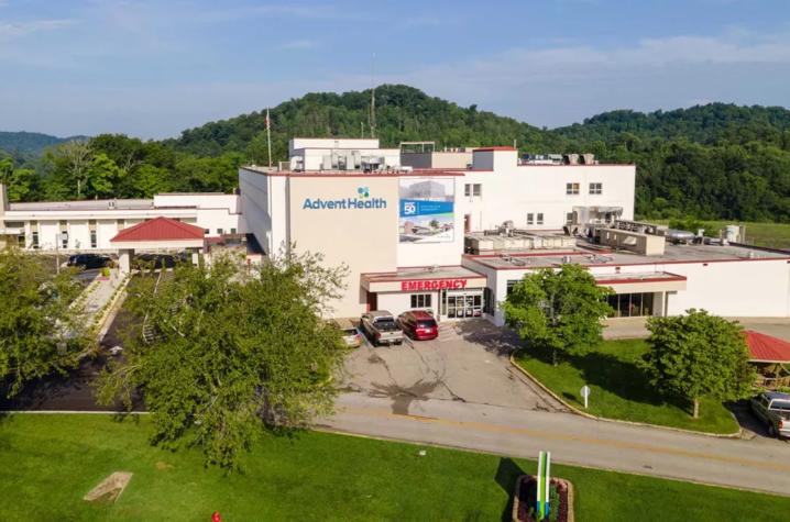 AdventHealth Manchester hospital facility aerial view showing the main building with Emergency entrance, surrounded by green trees and hills. Aerial view of a large light-colored medical building complex with multiple entrances, parked vehicles and ambulance bays, surrounded by green trees and rolling hills under a clear sky.