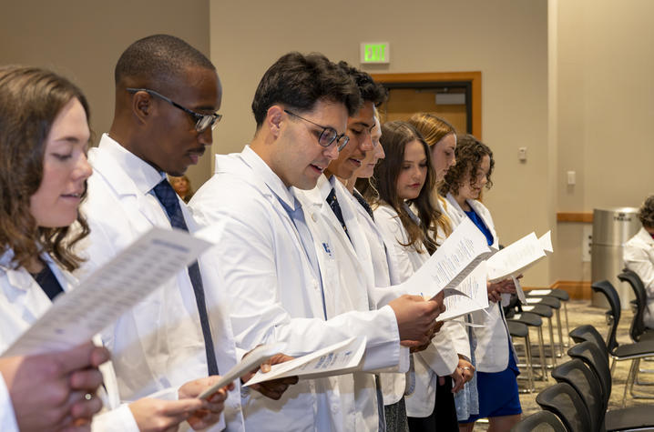 A group of people wearing white lab coats stand in a line indoors, holding printed programs or booklets and reading from them during a ceremony.