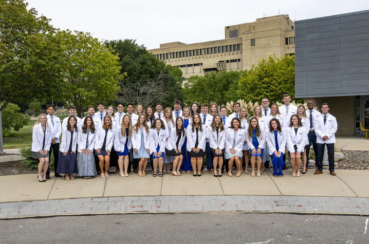 A large group of people wearing white lab coats pose for a group photo outdoors on a paved area in front of a campus building.