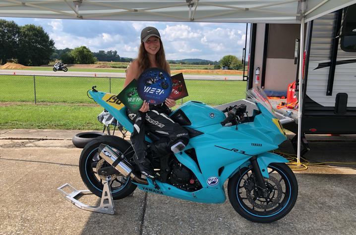 kelsey markle standing behind her motorcycle, smiling while holding three awards.