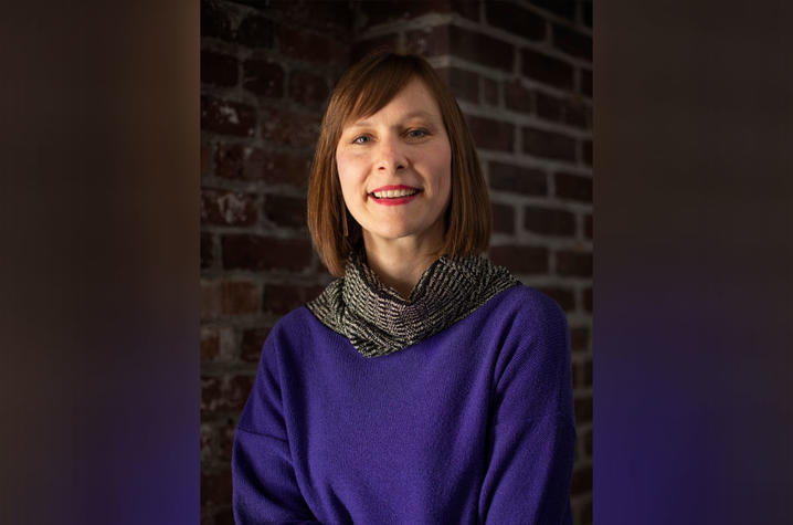 Headshot of a person with short brown hair wearing a purple sweater and patterned scarf, seated in front of a dark brick wall background.