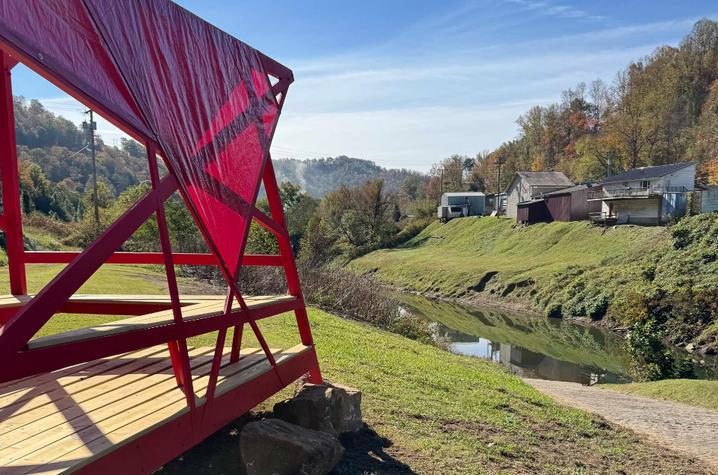A bright red geometric pavilion stands beside a narrow creek on a sunny day. The structure overlooks grassy banks, a walking path and a small cluster of homes, with wooded hills and a clear blue sky in the background.