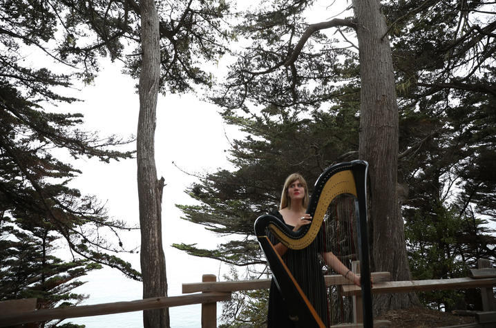 White woman wearing black dress standing outdoors with a harp.