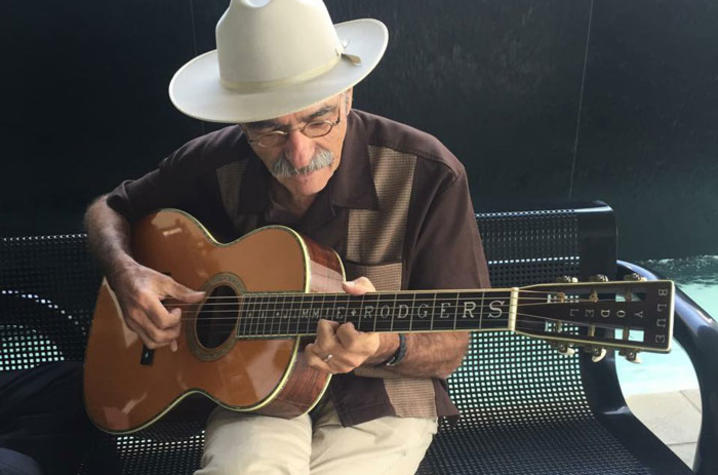 A man in a brown shirt and khaki pants sits on a bench playing guitar