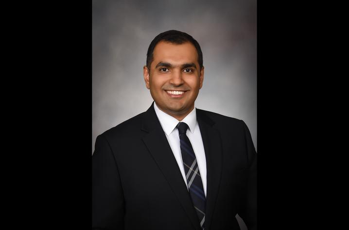 A headshot of Dr. Malik Khan who is wearing a black suit and black tie against a grey background.