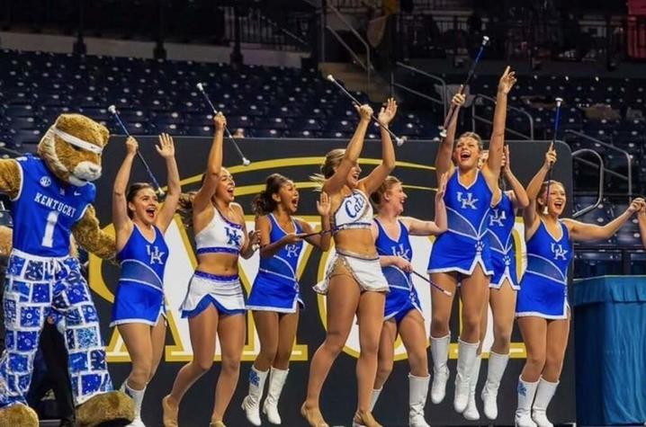 University mascot and dance team perform on a basketball court, raising their arms during a routine in an arena.