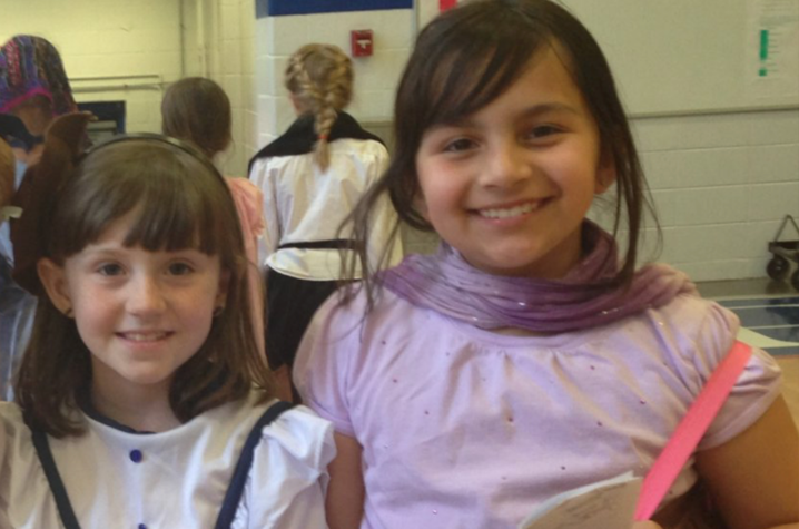 Two children standing together indoors at an event, wearing costume-style outfits, with other attendees visible in the background.