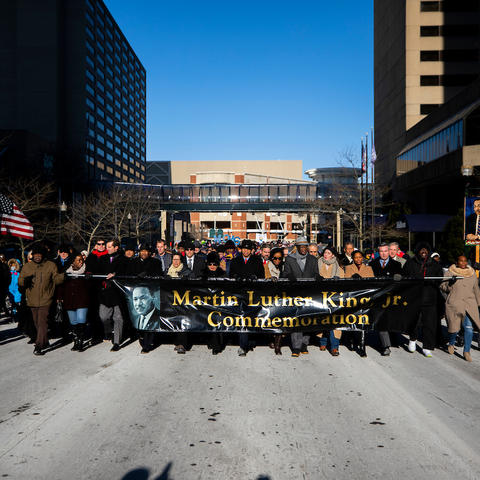 Large group marching on a snowy city street, holding a banner that reads “Martin Luther King Jr. Commemoration.”