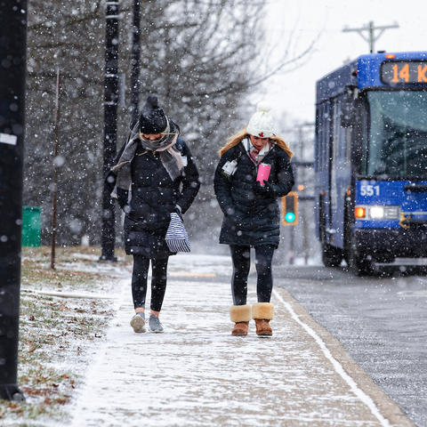 two students in the snow walking on a sidewalk next to a busy road