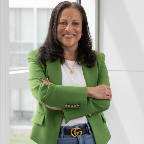 Image of Dr. Maria Braman, smiling and wearing a green blazer in front of a wall of windows