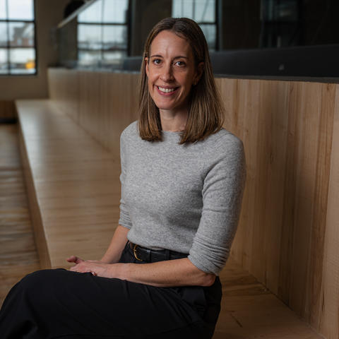 A person with shoulder‑length brown hair sits on wooden bleacher-style seating in a modern, spacious room with large industrial-style windows. They are wearing a light gray long-sleeve top and a dark skirt or pants, with hands resting in their lap. 