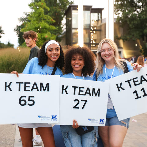 Three people hold signs reading “K TEAM 65,” “K TEAM 27” and “K TEAM 11” while standing outdoors on campus during a university event, with other attendees visible in the background.