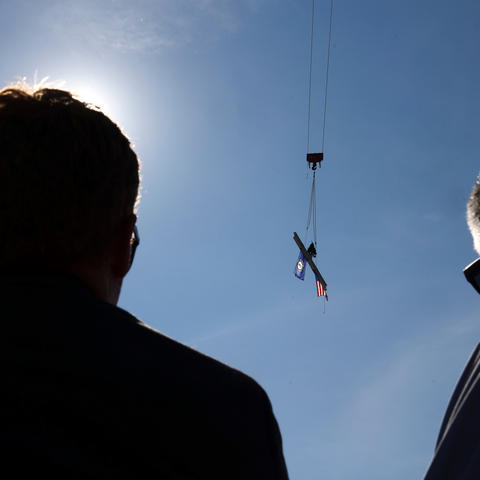 Two people in silhouette watch a crane lift a steel beam carrying an American flag and a Kentucky flag against a clear sky.