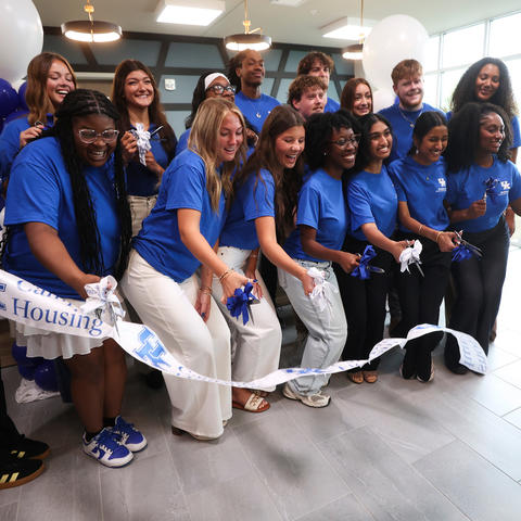 Group of students and university leaders in blue shirts gather indoors for a ribbon-cutting ceremony, holding ceremonial scissors and ribbon beneath white balloons.