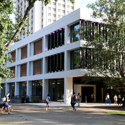 Students walk along a shaded campus pathway outside a modern academic building surrounded by large trees.