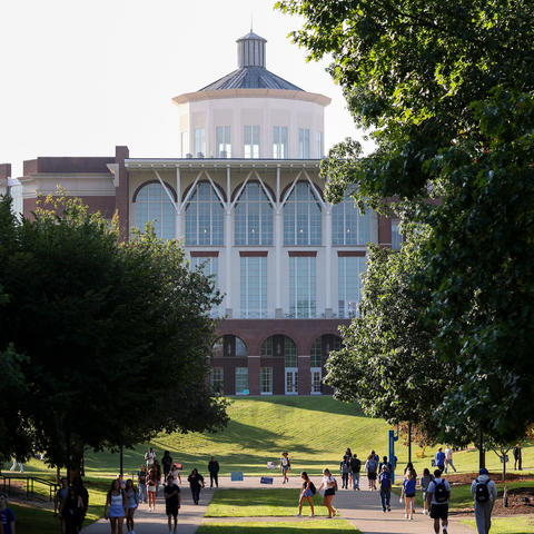 A large academic building stands at the end of a tree-lined campus walkway. Students walk along the path and gather on grassy areas beneath the trees on a sunny day.