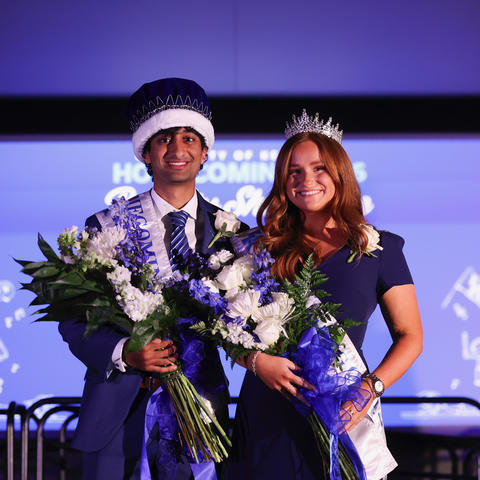 Two people wearing Homecoming crowns and sashes pose together while holding bouquets of flowers.
