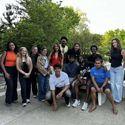 A group of young adults pose outdoors, standing and sitting around a bench with leafy trees behind them.