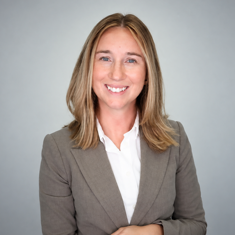 A smiling woman with shoulder-length blonde hair wears a gray blazer and white shirt against a soft gray background.