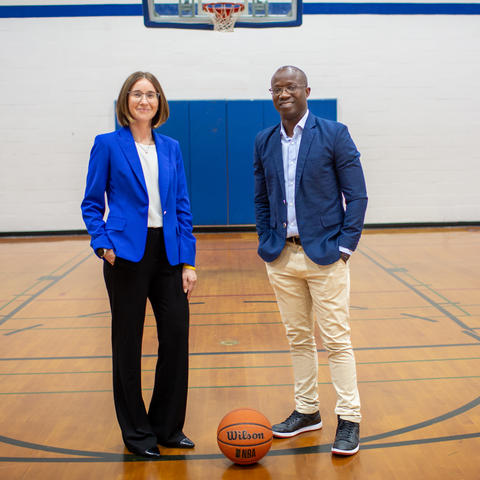  Two people stand on a gym basketball court beneath a hoop, with a basketball on the floor between them.