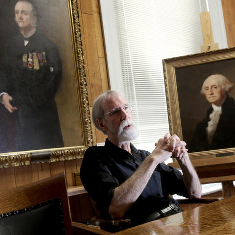 photo of Barry Bauman seated in front of portraits of Lucius Fairchild and George Washington by artists by John Singer Sargent and Thomas Sully