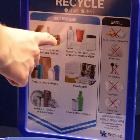 A person points to a recycling guide on a blue bin showing what items can and cannot be recycled at the University of Kentucky.