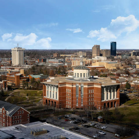 Aerial view of the UK campus with a large red‑brick library building in the foreground, surrounding academic buildings, tree‑lined walkways, parking areas, and a distant city skyline under a blue sky with scattered clouds.