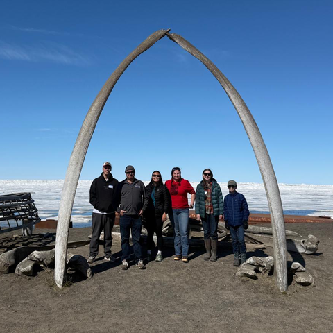 Six people stand beneath a large arch formed by two whale bones on a dark shoreline, with wooden boats and a vast expanse of sea ice beneath a clear blue sky in the background.