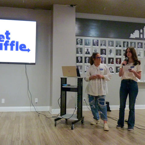 Two presenters stand at the front of a room giving a presentation beside a large screen displaying the logo “Sheet Shuffle.” One person speaks into a microphone while the other stands near a laptop on a cart. A photo wall is visible in the background.