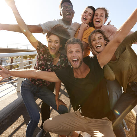 Six diverse young adults with big smiles and outstretched arms are huddled for a group photo. They are outside on a sunny day and appear to be standing on a pedestrian bridge. 