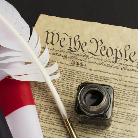 staged photo of copy of Constitution with a quill and bottle of ink and American flag beside of it