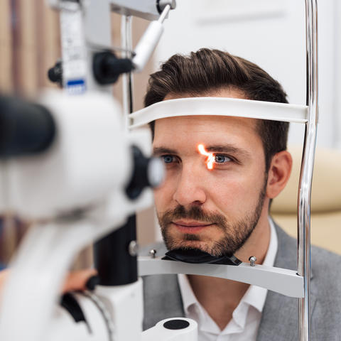 Photo of a young white man with dark hair and a beard getting his eyes examined.