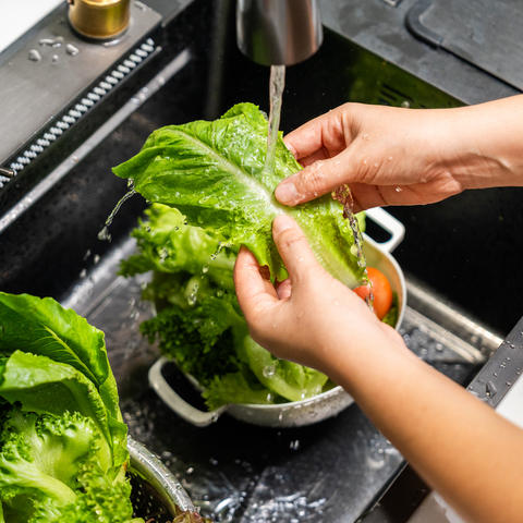 Hands rinse romaine lettuce under running water in a kitchen sink filled with fresh vegetables, including broccoli and leafy greens.