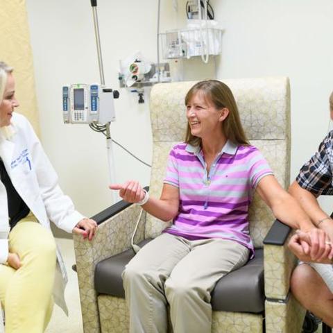 A young nurse in a white coat sits beside a middle-aged white woman in a hospital chair; her husband is sitting beside her and holding her hand. 