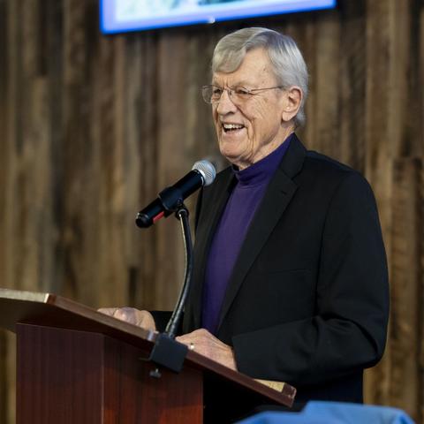 A person stands at a wooden podium speaking into a microphone, wearing a dark blazer and purple shirt, with a rustic wooden wall and a screen in the background.