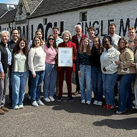 A group of about 20 people stand outdoors in front of a white stone building with large black painted lettering on the wall. One person in the center holds a framed certificate while the group gathers around for a photo.