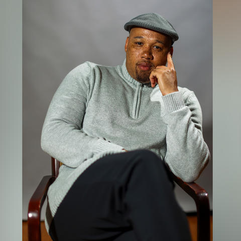 Black man wearing a grey sweater and black pants seated in a chair.